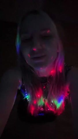 A woman is recording a video of herself under disco lights. The lights are red, green, and blue. She is wearing a black top and necklace. She briefly touches her hair with her hand. The background is dark. Music plays.