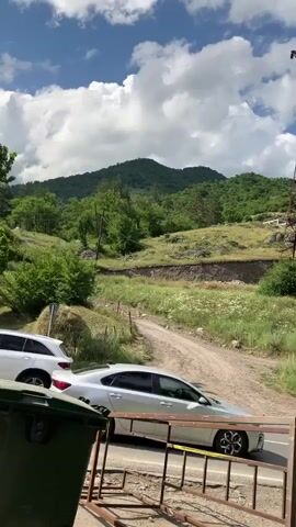 This video captures a scene in Dilijan, Armenia, under a bright, cloudy sky, suggesting a daytime setting with potentially intermittent sun. The foreground is partially obscured by a large, green trash bin and a rusty metal barrier. Beyond this, a dirt road winds its way up a grassy, hilly landscape dotted with trees.

A prominent feature of the scene is a tall, lattice-style electricity pylon, which is crossed by overhead power lines. These lines stretch across the frame, contributing to the sense of rural infrastructure. In the middle ground, a dark sedan is visible driving on a paved road that runs parallel to the dirt road. The car appears to be moving away from the camera.

The background is dominated by rolling, green hills covered in dense foliage. The landscape suggests a natural, perhaps somewhat undeveloped, area. There are no visible people or explicit text in the video, but the presence of the car and the electricity pylon indicates human activity and infrastructure within this natural setting. The overall impression is one of a serene, yet functional, rural environment.