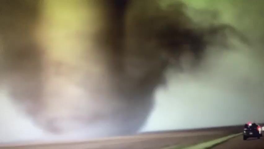 A tornado is seen on the right side of the screen, with a dark funnel cloud reaching down to the ground. A highway is visible in the foreground, with a black car driving on the right side of the road, away from the tornado. The sky is cloudy and appears to be a yellowish-green color. The overall scene depicts a natural disaster in progress, with the tornado posing a significant threat to the car on the highway.