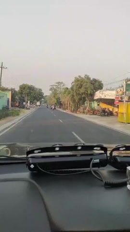 The video is a first-person perspective from inside a car, looking out through the windshield. The car is driving on a paved road, which appears to be a two-lane highway or main road. On either side of the road, there are trees, vegetation, and some buildings that look like houses or small shops. There are other vehicles on the road, including motorcycles and cars. The sky is a light color, suggesting it might be early morning or late afternoon. In the foreground, on the dashboard, there are some accessories, including what looks like a car air freshener and some electronic devices. The video shows a typical scene of driving through a populated area, possibly a town or village, with a clear sky and moderate traffic.