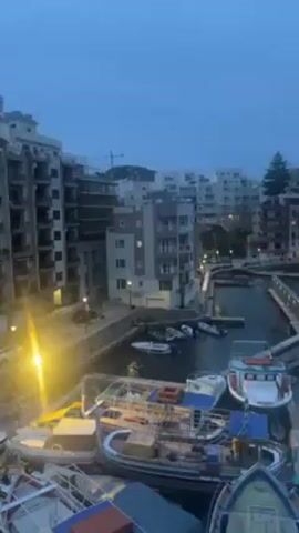 The video shows a harbor and the adjacent town, with the scene captured at dusk or early morning under a cloudy sky. Several boats are docked in the harbor, which is surrounded by buildings. Street lights and building lights are visible, reflecting on the water's surface, indicating that it's likely after sunset or before sunrise. A construction crane can be seen in the background, suggesting ongoing construction in the area.