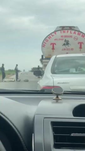 This media file depicts a traffic scene on a road in Kwali, Nigeria. The perspective is from inside a vehicle, looking forward through the windshield.

The primary subjects are several individuals standing on the road. Two of them, appearing to be men in dark uniforms or clothing, are positioned near the left side of the road. One is facing away from the camera, while the other is looking towards the distance. They are standing near what appears to be a makeshift barrier or roadblock, with a sign that reads "DIVERSION." A third individual, also in dark attire, is further down the road, seemingly engaged in some activity or directing traffic.

Several vehicles are visible. In the foreground, a white Toyota Corolla is stopped. To the left of the Corolla and across the road, a large tanker truck is present. The tanker has prominent red lettering on its side. The visible text on the tanker includes "HIGHLY INFLAM," "NO SMOKING," "40,000," and "JJN 833 XD." There is also a stylized logo of a horse and rider. Further down the road, other vehicles and individuals are faintly visible, suggesting a traffic slowdown or blockage.

The scene appears to be taking place during daylight hours, under an overcast or cloudy sky, indicating possibly dull weather. The road surface shows signs of wear and tear. The overall atmosphere suggests a temporary obstruction or traffic management situation, with the presence of the "DIVERSION" sign and individuals managing the flow of vehicles. The presence of uniformed individuals could indicate some form of official presence or authority enforcing the diversion.