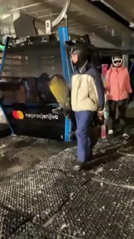 Several individuals exit a modern ski lift gondola in what appears to be a ski resort environment. They are dressed in winter sports attire, including jackets, helmets, and goggles, and carry skis or a snowboard. The gondola features modern branding and design, with dark paneling and blue accents. The ground is covered in a mix of snow and a dark, rubberized mat, providing traction. The environment suggests an indoor loading or unloading area for the ski lift, likely within a building or covered structure.