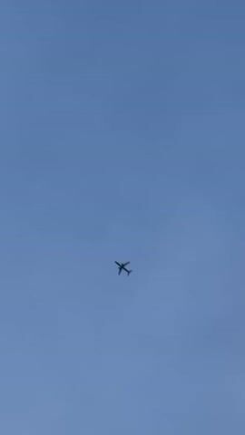 A small airplane is visible flying across a clear blue sky. The plane is dark in color and is seen from below, with its wings and tail clearly discernible. The camera appears to be stationary, and the plane is moving from left to right in the frame. The shot is focused on the airplane, with the background being an uninterrupted expanse of sky. There are no other objects or people visible in the frame.