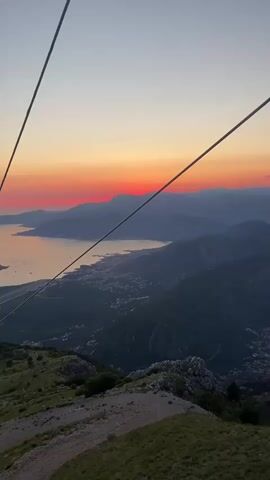A vertical video captures a panoramic view from a cable car descending a mountain at sunset. The sky is a gradient of colors, from a soft blue at the top to vibrant orange and red near the horizon. The sun itself is a bright red disc peeking from behind a distant mountain range. Below, a body of water, likely a bay or sea, stretches out, reflecting the sunset colors. A coastal town with buildings and lights is visible along the shoreline. Mountains rise on either side of the water, covered in dark green vegetation. The foreground features the rocky, grassy slope of the mountain where the cable car is located. Two thick cables of the cable car system run diagonally across the frame from the top left to the bottom right. A single red cable car cabin is visible moving downwards on the right-hand cable, with its door slightly ajar. The overall impression is one of breathtaking natural beauty and scenic travel.