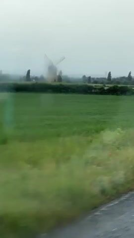 The video captures a view from a moving vehicle, looking out across a green field. In the distance, a windmill with rotating blades is visible. The sky is overcast, suggesting a cloudy or rainy day. The foreground shows blurred greenery, indicating movement.