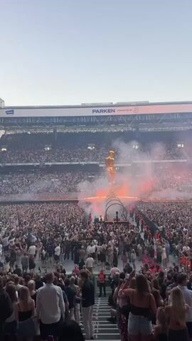 This video shows a large crowd of people in a stadium. A tall, golden statue stands on a stage in the center of the stadium, surrounded by smoke and orange lights. The crowd is cheering and holding up their phones. The stadium is mostly full, with people in the stands and on the field. The sky above is clear and pale. The word "PARKEN" is visible on the stadium roof.