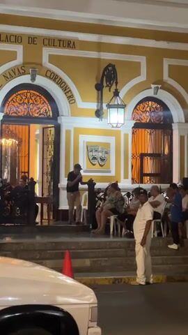 A group of people are gathered outside a yellow building at night. A man is standing on steps in front of the building, holding a microphone and speaking. Other people are sitting on chairs in front of the building, facing the speaker. There are also some people standing. The building has arches and windows, and the words "CUENTAS CORDENERO" and "DE CULTURA" are visible above the entrance. The scene appears to be an outdoor performance or event.