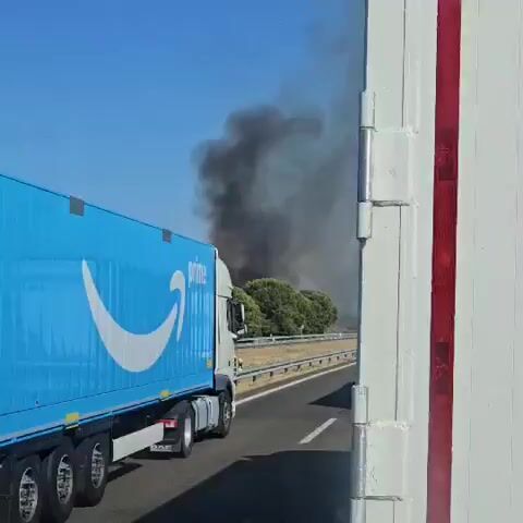 An Amazon Prime truck drives on a highway. In the background, a plume of thick black smoke rises into a clear blue sky, indicating a fire. The truck is blue with the Amazon Prime logo on its side. The truck is traveling to the left of the frame. The smoke is visible behind a line of trees and brush. The right side of the frame shows the white interior of another vehicle.