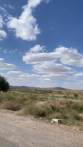 The video depicts a roadside view from a moving vehicle. Initially, a small tree and a kilometer marker are visible in a field under a partly cloudy blue sky. As the video progresses, the view shifts to show more of the landscape, featuring a utility pole, overhead power lines, and guardrails along the road. The road appears to be paved, and the surrounding terrain is a mix of sparse vegetation and open fields, with distant hills visible on the horizon.