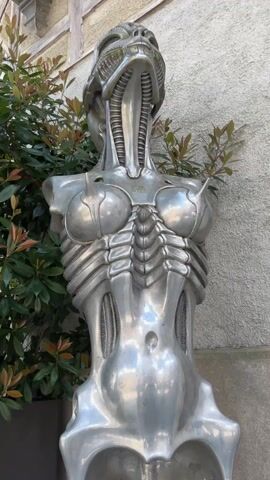 The video shows an outdoor area near the HR Giger Museum in Gruyeres, Switzerland. It begins with a close-up of a metallic sculpture of a female torso with biomechanical features. The camera then pans out to show a sign pointing to the "Museum Shop" with a poster advertising an exhibition of HR Giger's photographs. Further panning reveals an outdoor seating area and the facade of a multi-story building with a combination of gray and yellow walls and a small tower. The sky is blue with scattered clouds, and there are trees and greenery around.