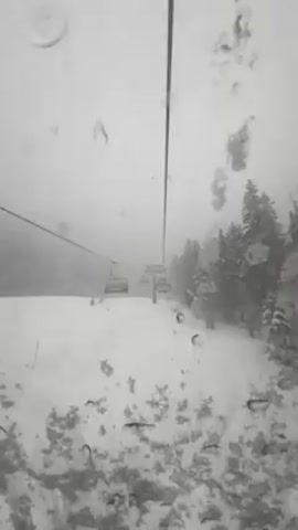 The video shows a view from a ski lift, looking down a snow-covered slope. A line of ski lift chairs extends into the distance. The sky is overcast and there are raindrops or condensation on the window. Evergreen trees line the right side of the slope. A skier can be seen briefly on the slope.