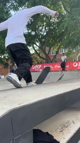 A video shows a person attempting to perform a trick on a skateboard. The person is standing on the side of a concrete skate park obstacle, holding their skateboard. They attempt to ollie onto the obstacle but miss and the skateboard slides out from under them. The person catches the skateboard and smiles before walking away. In the background, there are green trees, a brick building, and a red banner with "FCP" written on it.