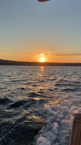The video shows a sunset over a body of water from the perspective of a moving boat. The sun is a bright orange orb, partially obscured by a few thin clouds near the horizon. The water is dark blue with gentle waves, reflecting the sunlight in a shimmering path. The boat's wake is visible, creating white foam behind. The sky is a gradient of light blue to orange as it approaches the horizon. A dark silhouette of land with some buildings is visible in the distance. The boat's wooden deck is partially visible at the bottom right corner of the frame.