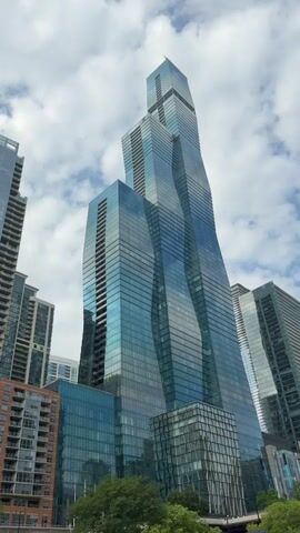 The video shows a low-angle shot of a modern, tall skyscraper with a distinctive twisted design. The building is predominantly made of glass with blue and teal tones. It is surrounded by other tall buildings, suggesting an urban environment. The sky above is partly cloudy. Green trees are visible at the bottom of the frame, hinting at a park or waterfront area. The text overlays indicate that the building features retail space and a parking garage, and that its name is a "name sake."