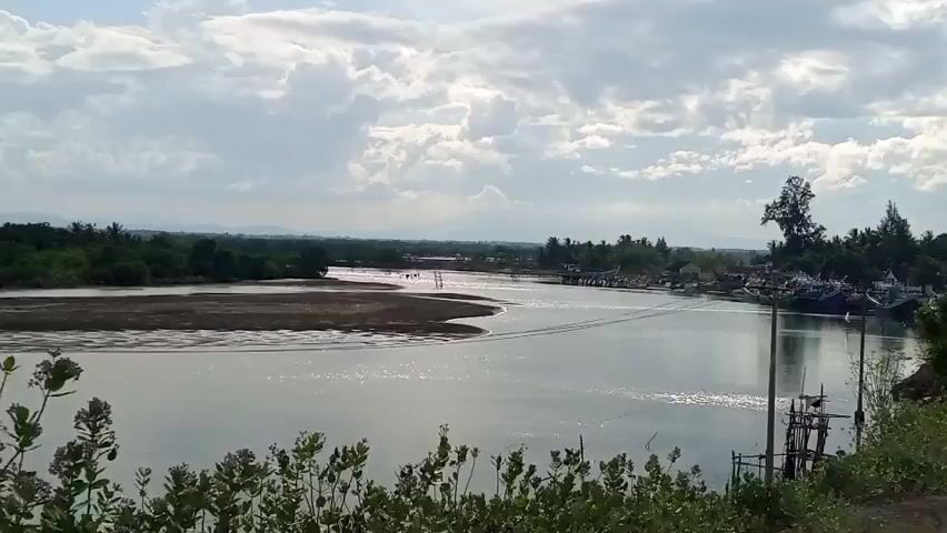 A wide shot captures a serene river with boats docked on the right side. The river flows from left to right, with a sandy bank on the left and lush greenery lining the shores. The sky is a mix of blue and white clouds, casting a soft light on the landscape. The water reflects the sky, creating a shimmering effect. A few trees are visible on the right bank, and a road runs alongside the river, with a motorcycle parked on it. The overall scene is peaceful and tranquil, evoking a sense of calm and natural beauty.