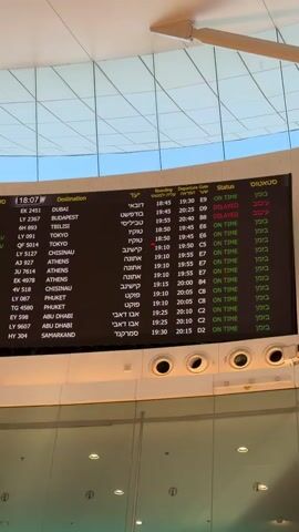 This is a close-up shot of a departures board at an airport. The screen displays flight information in both English and Hebrew, with columns for Destination, Flight Number, Boarding Time, Departure Gate, and Status. The top left corner shows the current time as 18:07.

The subjects are the flight information displayed on the screen, which functions as a central object in the scene. The setting is clearly an airport terminal, indicated by the presence of the departures board, the modern architecture of the ceiling with its angled panels, and the metallic fixtures below the screen.

The activity taking place is the display of real-time flight information, showing destinations like Dubai, Tbilisi, Tokyo, Athens, and Abu Dhabi. Some flights are marked as "ON TIME" in green, while others are "DELAYED" in red. The time of day is indicated as 18:07 on the board, suggesting it is early evening. There are no visible people in the frame, but the presence of a finger in the lower left corner of some frames suggests someone is interacting with or capturing the image. The overall emotion conveyed is one of travel and the anticipation or uncertainty associated with flight schedules.

Notable details include the distinct red markings for delayed flights, the use of two languages on the board, and the specific flight times and gate assignments. The architecture of the airport is modern and sleek.