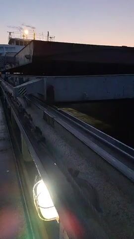 The video opens on a close-up, low-angle shot of a metal railing with a light fixture attached. The railing runs along the side of a barge or large vessel. In the background, the dark silhouette of the vessel's deck is visible, as well as a building structure with lights on top. The sky above is a gradient of dusky blue and purple, indicating dawn or dusk. A chain is visible secured along the top of the railing. The camera appears to be moving, as evidenced by the slight blur in the foreground. Two figures, appearing to be people, are dimly visible walking on the deck in the distance, on the left side of the frame. The overall atmosphere is industrial and maritime.