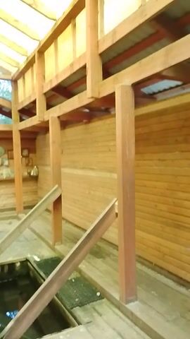 The video shows an interior view of a wooden structure. The camera is tilted and appears to be moving, providing a dynamic perspective of the exposed wooden beams and ceiling. The ceiling is made of horizontal wooden planks. Several wooden beams are visible, forming a supporting structure for the roof. Some of these beams are angled, providing structural support. In the background, there is a wall adorned with numerous framed pictures and what appear to be religious icons. A doorway or opening is visible on the right side, leading to a darker area. The floor is also made of wooden planks. The environment suggests a rustic or traditional architectural style.