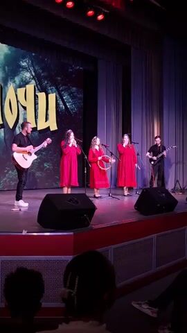 A stage performance featuring five people is underway. The stage is bordered by a deep red platform, and a large screen behind the performers shows an image of a forest under the word "HOYU." Three women are centrally located, all wearing red dresses, each holding a microphone. The woman in the center is also holding a tambourine. To the left of the women, a man in a black shirt and white pants is playing an acoustic guitar. To the right, another man dressed in black is playing an electric guitar. The stage is lit by red spotlights above, with two large black speakers positioned in front of the stage. Below the stage is a partially visible audience.