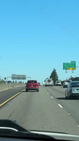 The media captures a view from inside a vehicle driving on a highway on a clear, sunny day. The scene is set in Ridgefield, United States, with a bright blue sky overhead and lush green trees lining the road. Traffic is moderate, with several cars visible ahead, including a red SUV and a white truck with a trailer. A green highway sign is visible on the right side of the road, indicating an exit for NE 179th St, Clark County Event Center, and Expo Center. The time of day appears to be late morning or early afternoon, given the bright sunlight and clear visibility. The overall mood of the scene is peaceful and ordinary, depicting a typical commute or road trip.