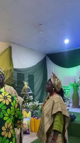 Two people in traditional African garb stand in a room decorated with green and white drapes. There is a table with gifts and flowers. One woman holds a picture of a dog.