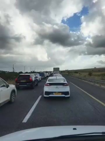 The video shows a line of vehicles in heavy traffic on a highway. The sky above is mostly cloudy with patches of blue. The view is from the interior of a car, looking forward at other cars that are either stopped or moving very slowly. The car directly in front is a white Hyundai Ioniq with a license plate reading "175 28 501". Other cars, including a black Volvo SUV, are visible in the line. The landscape alongside the road includes grassy fields and occasional trees. Light poles are visible along the highway. The road markings include solid yellow and white lines.