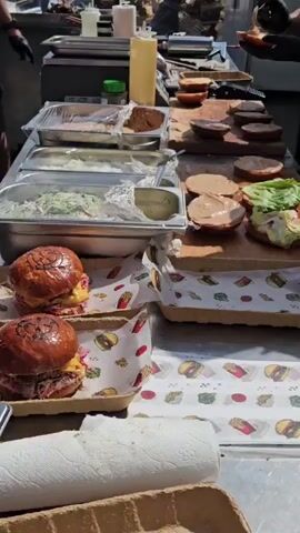 The video shows two people preparing burgers at an outdoor food stall. In the foreground, there are two assembled burgers on trays lined with paper. A table in the middle is covered with burger buns, some with sauce and lettuce, and ingredients for burgers. In the background, there are various containers of food, condiments, and kitchen equipment. The two individuals, wearing black gloves and aprons, are actively assembling burgers. One person is placing a burger patty on a bun, while the other is handling ingredients. The environment appears to be sunny with trees and a large white refrigerator visible in the background.