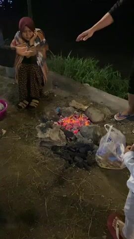 The video opens with a close-up of a campfire with glowing embers. In the background, a woman wearing a hijab and a patterned skirt is sitting and appears to be crying or upset. Two other individuals are partially visible, one on the left and one on the right, with their hands extended. The scene then pans out to reveal a campsite at night. Numerous orange and pink tents are set up, and people are gathered around the campfire and near the tents. Some individuals are sitting on the ground, and there are plates of food and drinks visible. Lamps illuminate the area, and a body of water is in the background. The overall atmosphere suggests a group camping trip or gathering at night.