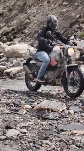 A person riding a motorcycle navigates a rocky, uneven terrain. The rider is wearing a helmet and dark clothing, with blue jeans. The motorcycle is a dark color. The environment is mountainous, with steep slopes covered in rocks and sparse vegetation. The road appears to be a dirt or gravel track, with sections of water or mud. The sky is partly cloudy, with some patches of blue. The video transitions to show wider mountain vistas, with more greenery and dramatic cloud formations.