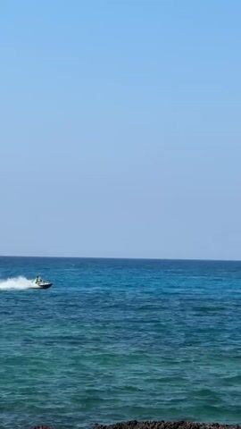 A jet ski with one person riding it is speeding across a bright blue ocean, creating a white spray of water behind it. The water is clear and the sky is a cloudless blue. In the foreground, there is a rocky shoreline.