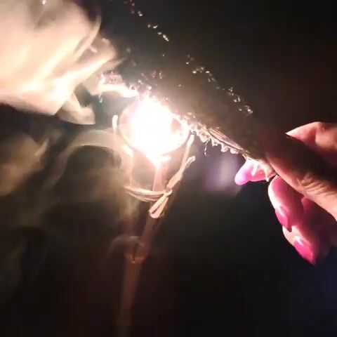 A hand with bright pink nails holds a burning sprig of dried herbs, likely sage, emitting smoke and sparks. The background is dark, suggesting it's nighttime or an unlit indoor space. A blurred, metallic object, possibly a small tripod or stand, is visible beneath the burning herbs, partially obscured by smoke. The burning process is captured in a series of short, repeating video frames.