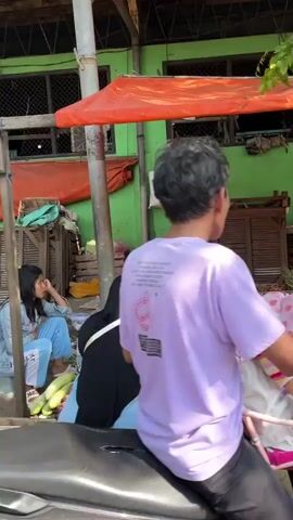 The video shows a street scene with several people and objects. In the foreground, a person wearing a purple t-shirt is sitting on a motorcycle, facing away from the camera. To the left, two young women are sitting on what appears to be a bench or ledge. One woman, wearing a light blue top and pants, is leaning forward with her arm resting on her knee. The other woman, wearing a black hijab and top, is partially visible. In front of the women, a pile of cucumbers is visible. Behind them, a green building with orange awnings is present, with some wooden structures and possibly market stalls. The environment appears to be outdoors, likely a market or busy street, with sunlight indicating daytime.