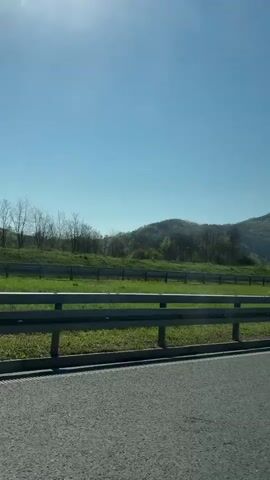 The video shows a view from a moving vehicle on a highway. A metal guardrail is visible along the side of the road. Beyond the guardrail is a green grassy area with scattered trees and a hill in the background. The sky is clear and blue with the sun visible in the upper left.