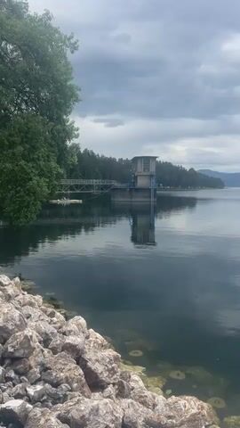 The video shows a calm lake surrounded by trees and mountains under an overcast sky. A concrete structure with a small building on top is connected to the shore by a narrow walkway. Rocks line the edge of the lake in the foreground. The water is dark and reflective, mirroring the sky and the structure. At the very end, faint ripples emanate from the lake surface.