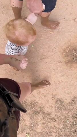 A group of people are standing in muddy ground. A baby with blonde hair is being held by its hands by two adults. The baby is wearing a striped shirt and is mostly obscured from view. The adults appear to be helping the baby walk through the mud. The ground is a light brown color and appears to be dirt or clay. There are some small green leaves scattered on the ground. The perspective of the camera is from a low angle, looking down at the scene.