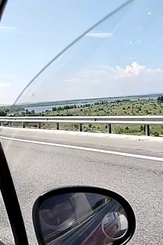 This is a first-person perspective video taken from inside a vehicle, looking out the passenger side window and at the side mirror. The vehicle is on a highway, indicated by the paved road, guardrail, and highway markers. The landscape outside the window is a rural scene with a body of water, green fields, and scattered trees under a clear blue sky with some clouds. The side mirror shows a reflection of the interior of the car and the road behind. The overall impression is of a leisurely drive on a sunny day.