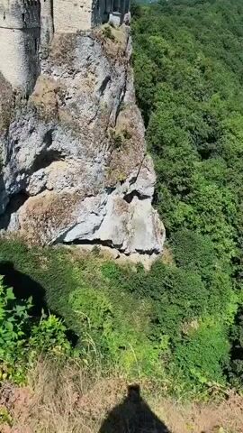 A drone captures a majestic castle perched precariously on a sheer rock cliff. The castle, made of stone, features a tall, white tower and several smaller turrets, with scaffolding visible on one side. A stone bridge connects to a lower section of the castle. The surrounding environment is lush and green, with a dense forest extending into the background under a clear blue sky. The camera pans and zooms to showcase the castle from various angles, emphasizing its dramatic location.