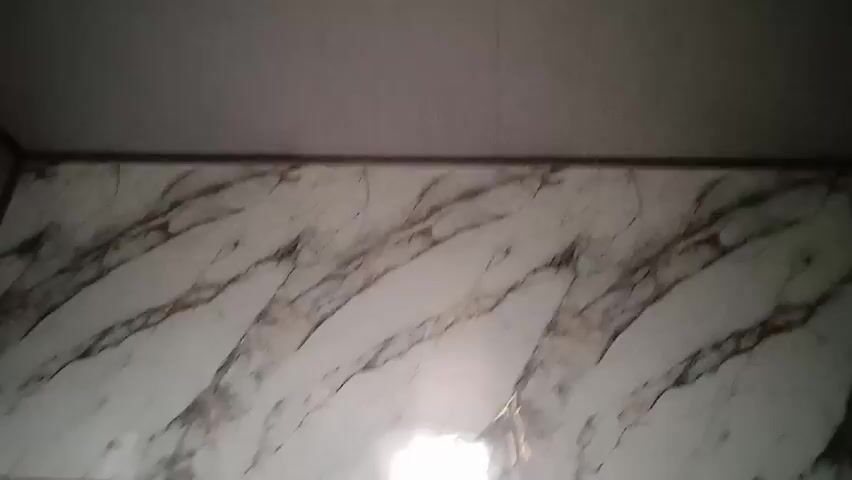The camera is pointed upward toward a section of tiled wall that appears to be in a bathroom or similar space. The tiles feature a white marble pattern with gray and brown veining. The tiles are framed by a dark trim at the top. Above the trim, there is a plain, light-colored ceiling.  There is no visible movement or people in the frame.