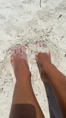 The video shows a top-down view of a person's bare feet partially buried in white sand. The feet are tanned, and there is sand on the toes and soles of the feet. The sand appears to be fine-grained. The lighting suggests it is daytime and sunny, likely at a beach. There are some small pieces of debris, possibly dried seaweed or twigs, scattered in the sand. The camera is stationary.