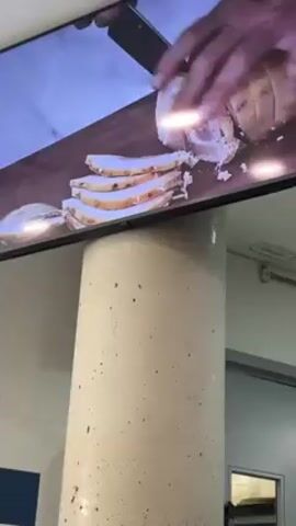 A close-up shot shows a flat-screen television mounted high up on a concrete pillar. The screen displays a close-up of hands slicing a roll of meat on a wooden cutting board. Four slices of meat lie to the left of the roll. The pillar, which is light gray with dark speckles, extends from the bottom of the frame and rises to the television. Behind the pillar, a tiled wall is visible, with blue and white tiles. To the right, there is an open doorway that leads into a dimly lit room. A security camera can be seen on the ceiling.