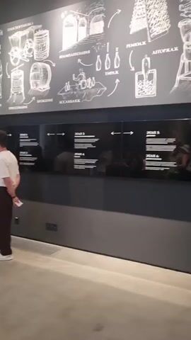 The video shows an interior view of a museum exhibit. On the wall, there is a large black and white diagram depicting a process, likely related to winemaking or beverage production, with various illustrations and text in a foreign language. Below this diagram is a horizontal black panel with white text arranged in columns, also in a foreign language, possibly detailing steps or information about the exhibit. Further down, a vertical panel displays a screen with a glowing white interface and a grid of circular elements below it. To the right of this, a wall is adorned with a collection of colorful vintage posters arranged in a staggered grid. In the foreground, a wooden workbench with a metal apparatus is visible. To the right of the workbench, another wooden stool-like structure supports a green piece of machinery. The overall environment appears to be dimly lit, with spotlights illuminating certain parts of the exhibit. The floor is a polished concrete or stone surface. No individuals are clearly identifiable in the video.