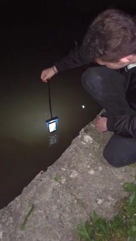 A dark, nighttime video shows a person, likely male, squatting or kneeling next to a body of dark water with a rugged concrete or stone bank. The individual is wearing a dark long-sleeved top and dark pants. They are holding a smartphone, enclosed in a black waterproof pouch, suspended by a lanyard. The phone's flashlight is turned on, emitting a strong beam of light into the murky water as it is slowly lowered. The flashlight illuminates a circular area underwater and creates reflections on the water's surface. The person appears to be observing the phone or the illuminated water. A bracelet is visible on their left wrist, and a small tattoo is on their right hand. The background is completely dark, indicating it is nighttime. The scene suggests an experiment or activity involving testing the phone's waterproof case or using its light to see underwater. No specific text is visible in the video frame.