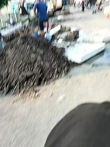 On an overcast day, a construction crew is working on a road, likely repairing or installing underground pipes. An excavator is positioned on the road, with its arm and bucket extended towards a freshly dug pit. Several workers are present, one standing near the pit and another in the pit itself. There are visible pipes and concrete slabs lying on the road, indicating the work being done involves underground utilities.  A pile of dirt sits near the excavation site. A warning sign with a graphic of a person falling and the words "AWAS ADA LUBANG GALIAN" (WARNING, THERE IS A HOLE) is placed on the side of the road. The surrounding environment includes buildings, trees, and power lines. A mosque with a green dome is visible in the background. Traffic is present on the road, indicating it is still open during construction.