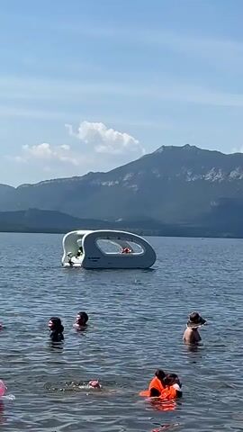 The video shows a lake on a clear day with a mountainous backdrop. In the foreground, several people are swimming and wading in the water. One person is wearing an orange life jacket. In the middle ground, a futuristic-looking white pod-like boat with windows floats on the water, with a person inside. The water is calm and blue, and the sky is a clear blue with a few white clouds. The mountains in the background are covered in trees and are rugged and imposing. The scene is peaceful and serene, suggesting a day of recreation and enjoyment.