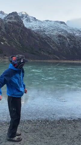 A young person, seen from the back, stands by the edge of an ice-covered lake, with majestic snow-capped mountains in the background. The person is wearing a blue and black jacket, black pants with a white logo on the left leg, and a patterned balaclava. They are standing on a gravelly shore, looking out at the expanse of the partly frozen, light-green lake. In the initial frame, the person is caught in the action of throwing something into the lake. The mountain slope has a few sparse trees. The sky is overcast.