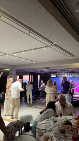 A band performs on a stage with a colorful backdrop in a dimly lit room. Several people are dancing in front of the stage, including a man in a white shirt and beige pants and a woman in a white dress. Other people are seated at tables covered with tablecloths, eating and drinking. One man, wearing a blue shirt and light-colored pants, is standing and facing the stage, appearing to sing along. Another man, seated at a table in the foreground, is wearing a white shirt. The room has a high ceiling with multiple lights, and large windows are visible in the background, showing a sunset or twilight sky. The tables are set with plates, glasses, and food.