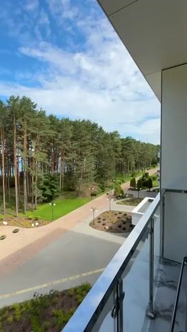 This media file presents a scenic view from a balcony overlooking a manicured park area with a dense pine forest bordering the scene. The sky is a vibrant blue with scattered white clouds, suggesting a pleasant day. The time of day appears to be late morning or early afternoon.

The foreground of the image is dominated by the balcony railing, made of metal and glass, indicating a modern building. Below the balcony, a paved walkway with a brick pattern is visible, lined with neatly trimmed green grass and low-lying shrubs. Several decorative round light fixtures are embedded in the landscaping, and lampposts with a modern design are strategically placed along the walkway.

Further into the scene, a circular paved area features a small, artistically shaped tree and several spherical decorative elements, adding to the park's aesthetic appeal. Beyond this, a long, winding path stretches towards the forest.

The main subjects in the scene are two individuals walking along the paved path. One person is seen pushing a stroller, while the other is walking with a red bag. Their presence suggests leisure activities or perhaps residents enjoying the outdoor space. The tall, straight trunks of the pine trees create a striking natural backdrop, with their green canopy extending across the horizon. The overall impression is one of tranquility and natural beauty, with a well-maintained environment designed for relaxation. The location context of Мядзельскі сельскі Савет, Belarus, suggests a rural or semi-rural setting within the country. No visible text is present in the media file.