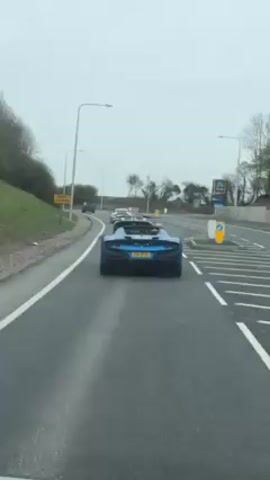 The video shows a line of luxury cars driving on a grey asphalt road. The camera is positioned in a car following behind them. The first car, closest to the camera, is a blue convertible with the license plate F8 DYU. Following closely behind are two more convertibles, and a white Porsche. There is grass on the left side of the road and trees, and on the right side of the road is a sign for Aldi. It is a cloudy day.