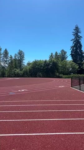 A red running track with white lines is shown under a clear blue sky with a few scattered clouds. Trees and a green field with goalposts are visible in the background. A black chain-link fence is in the foreground. The video is a stationary shot panning slightly to the right.